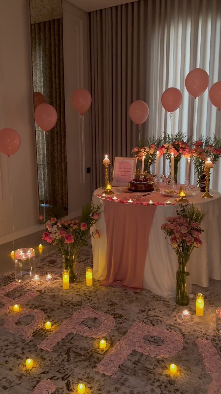 Romantic dinner table with pink balloons, candles, and rose petals on the floor.