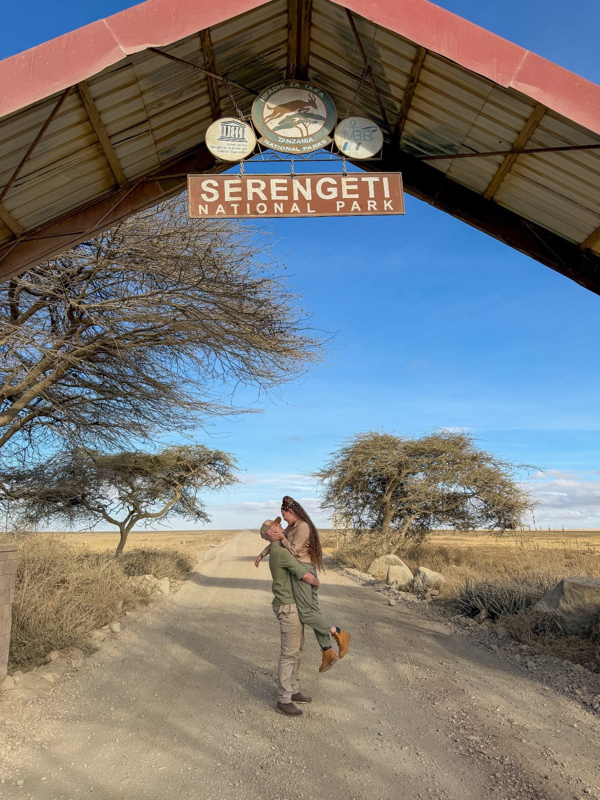 Couple kissing under the Serengeti National Park entrance sign on a dirt road.