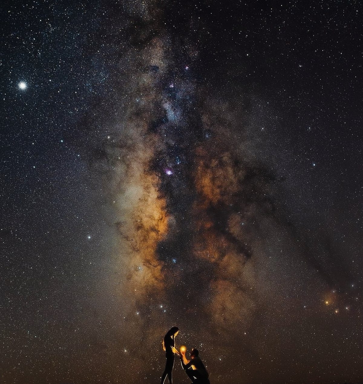 Silhouetted couple standing beneath a brilliant, starry Milky Way galaxy at night.