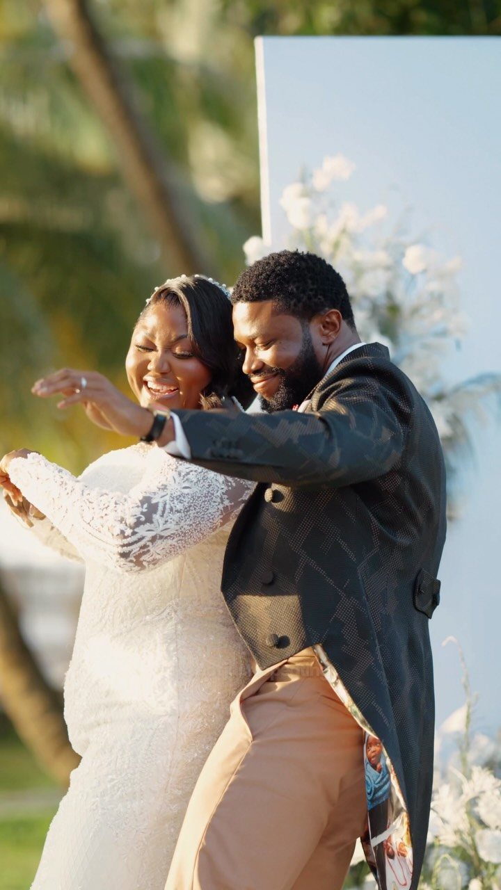 Smiling couple in wedding attire embracing outdoors in front of a floral backdrop