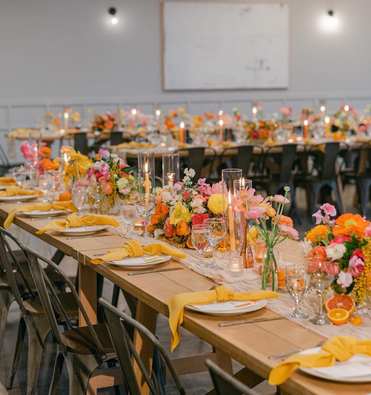 Wedding table setting with yellow napkins, bright floral centerpieces, and sliced citrus fruits.