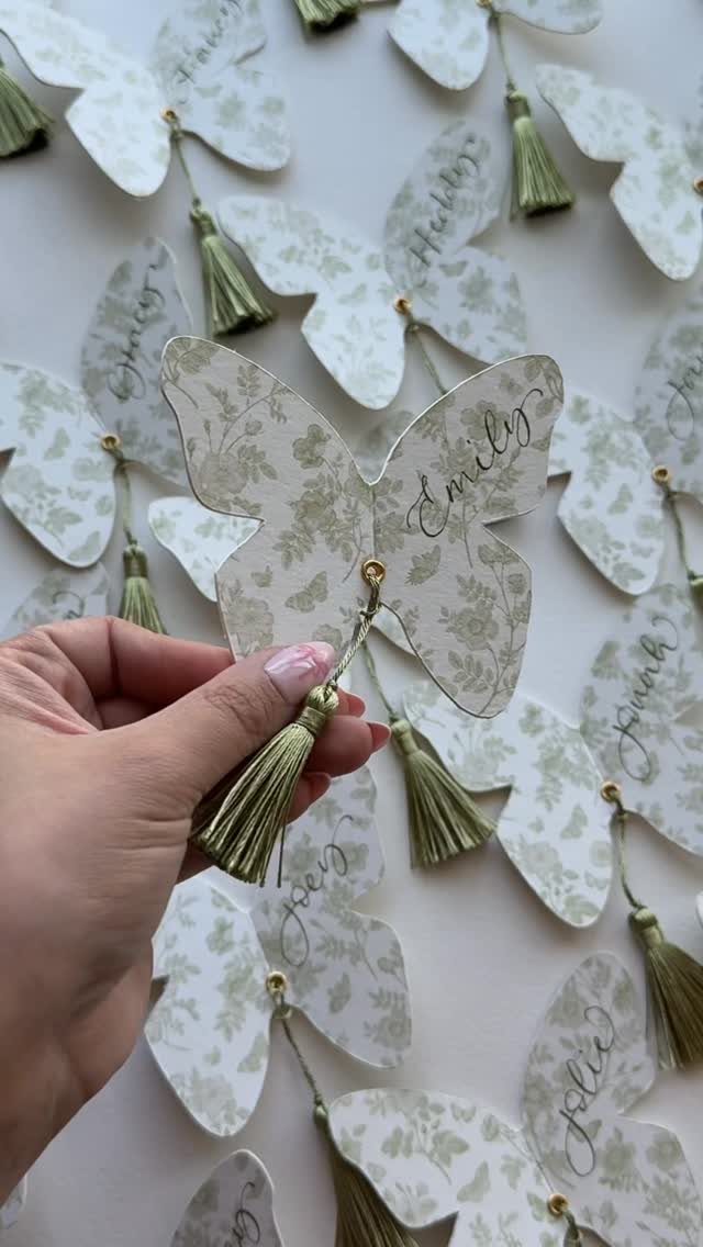 Butterfly-shaped place cards with green floral patterns and tassels held by a hand.