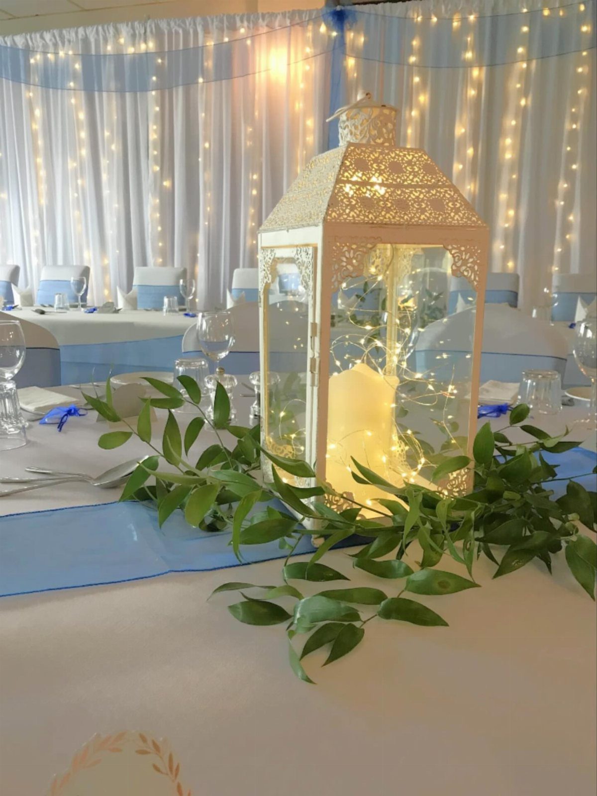 White decorative lantern with fairy lights and greenery on a wedding table setting