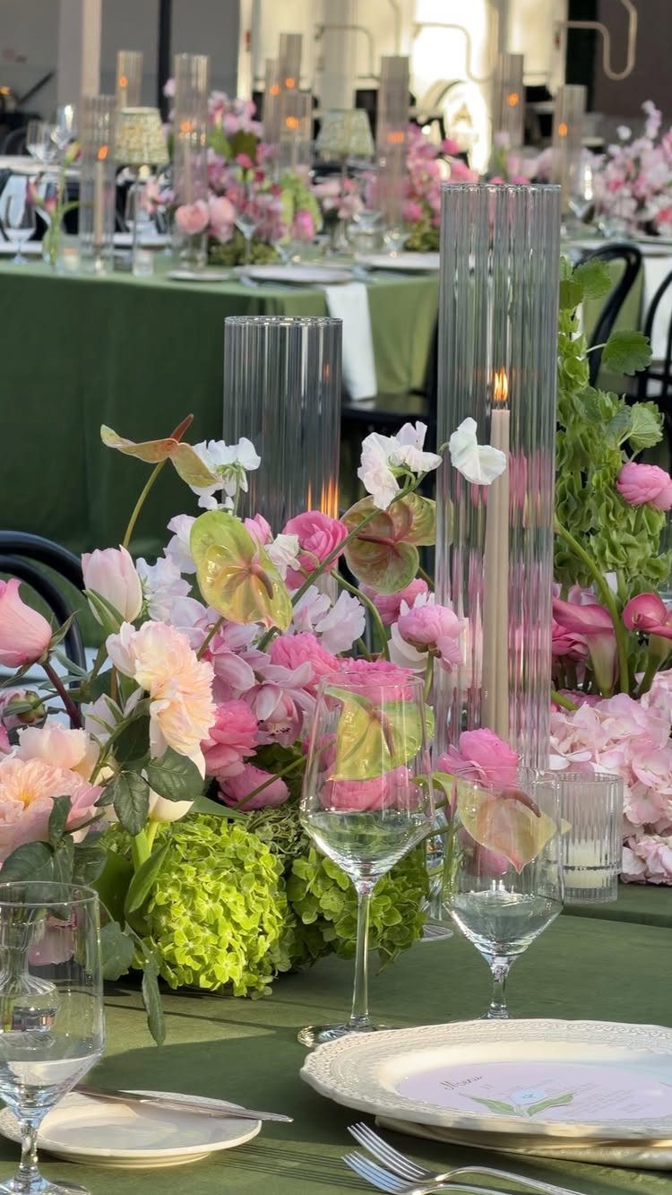 Wedding table setting with green linens, pink floral centerpieces, and tall glass candles.