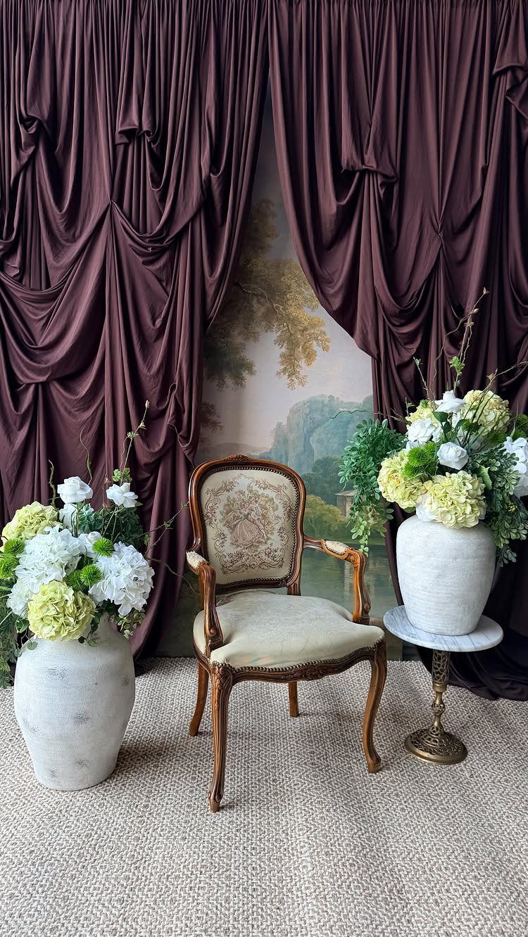 Vintage armchair with white floral arrangements and burgundy curtains in a lounge setting.