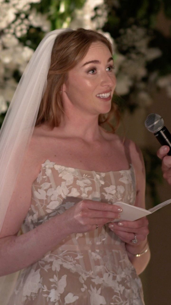 Bride in a floral lace dress and veil reading vows into a microphone
