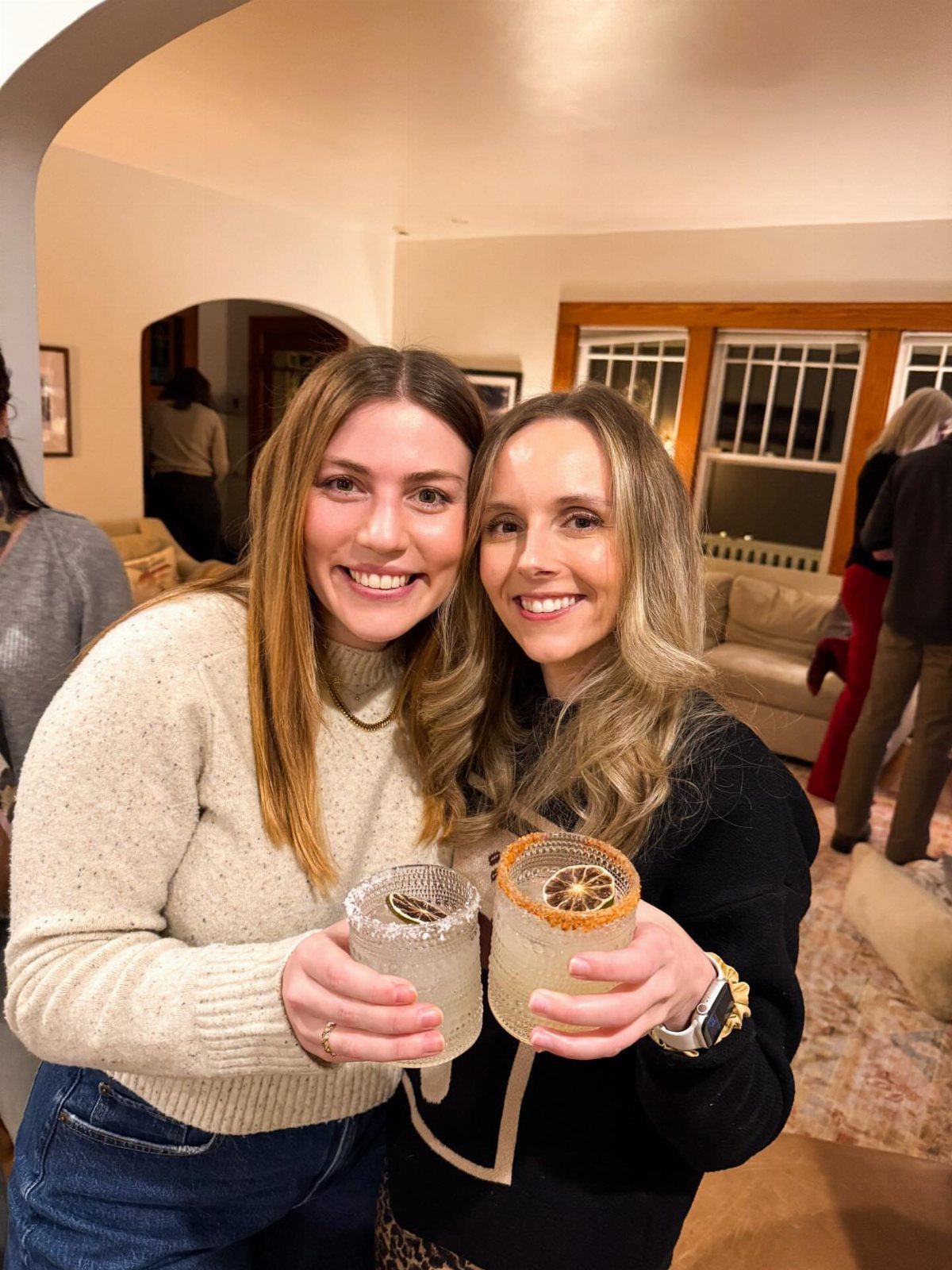 Two women smiling and holding cocktails in a home living room setting.
