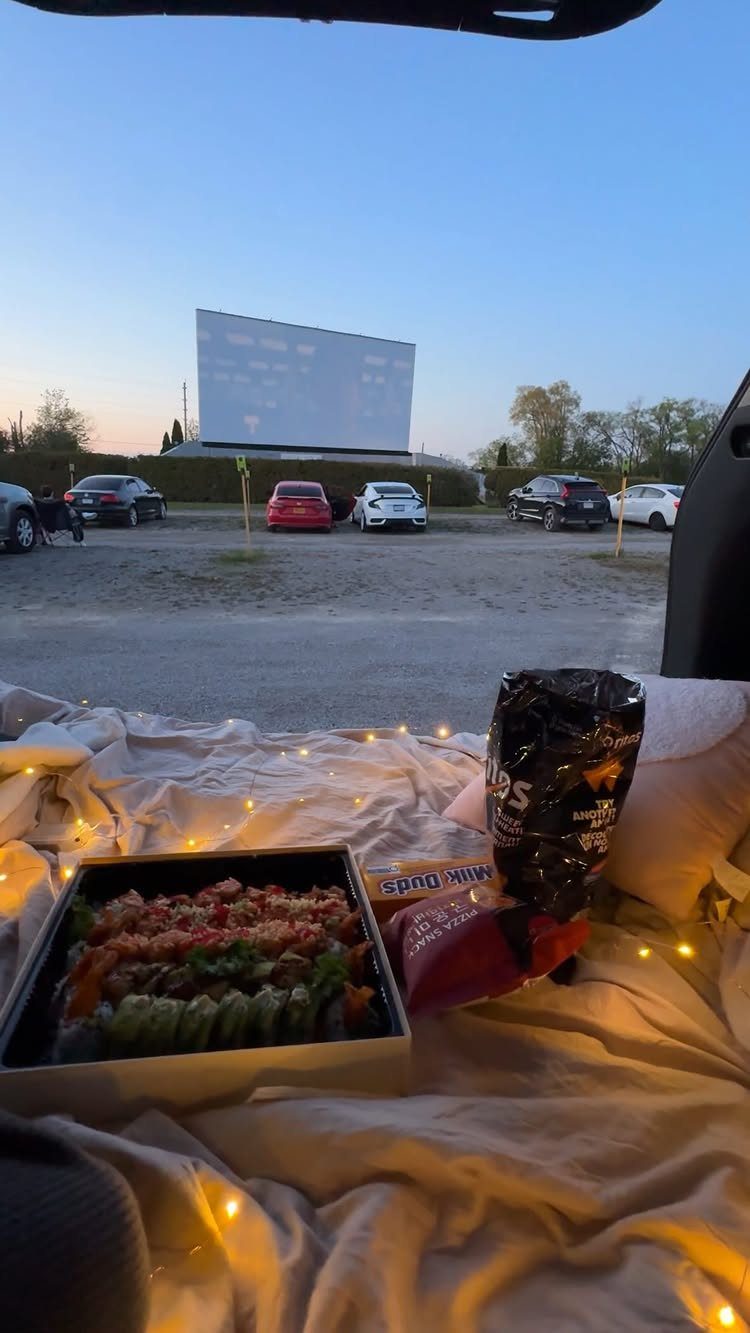 Drive-in movie screen viewed from a car interior with blankets, sushi, and fairy lights.