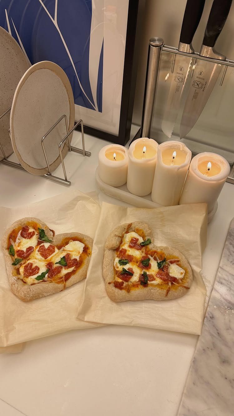 Two heart-shaped pizzas on parchment paper next to four lit white pillar candles.