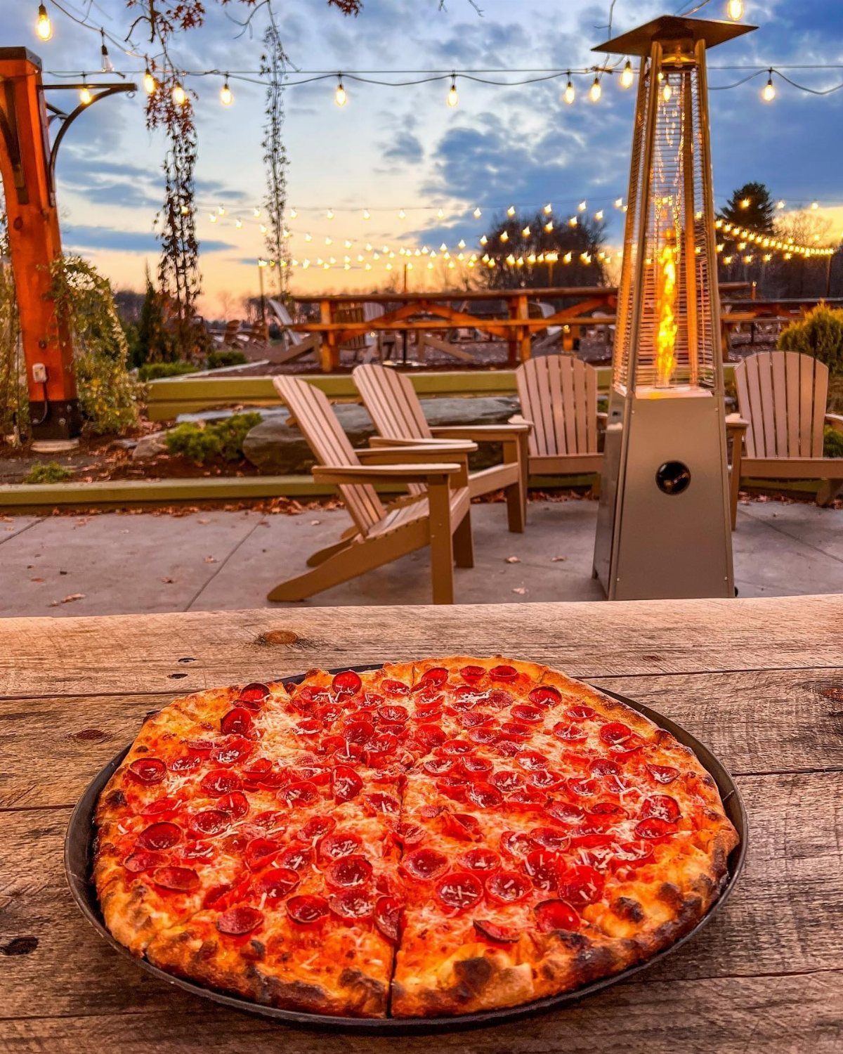 Pepperoni pizza on a wooden table with a patio heater and string lights.