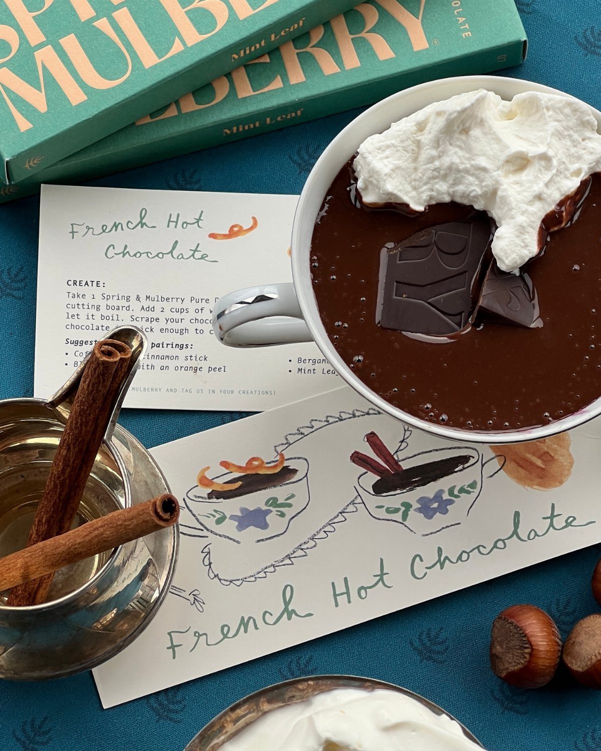 French hot chocolate with whipped cream, cinnamon sticks, and recipe cards on blue.