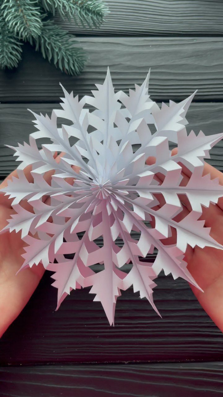 White 3D paper snowflake held by two hands over a dark wooden background.