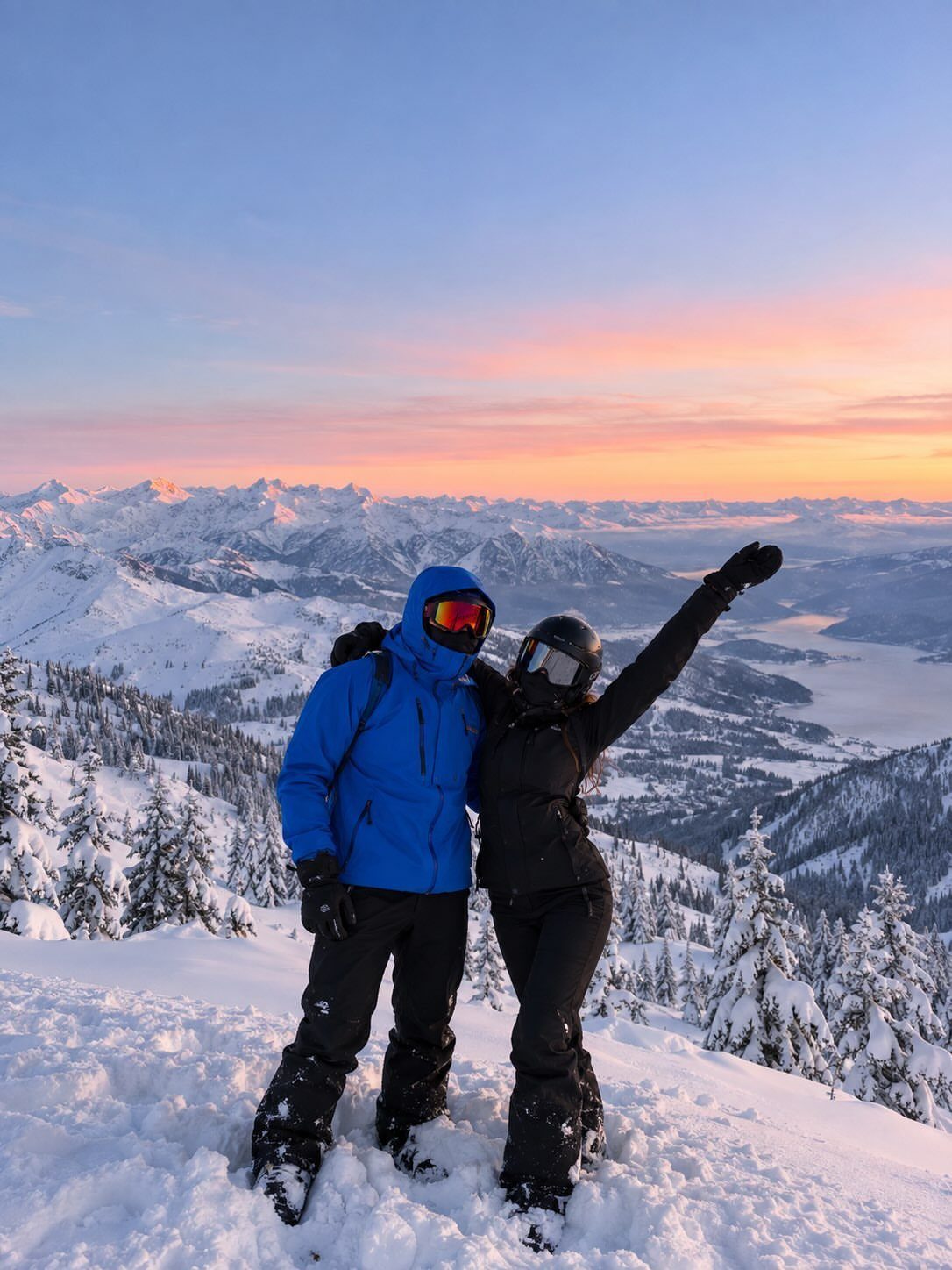 Couple in winter clothing posing on a snowy mountain peak at sunset.