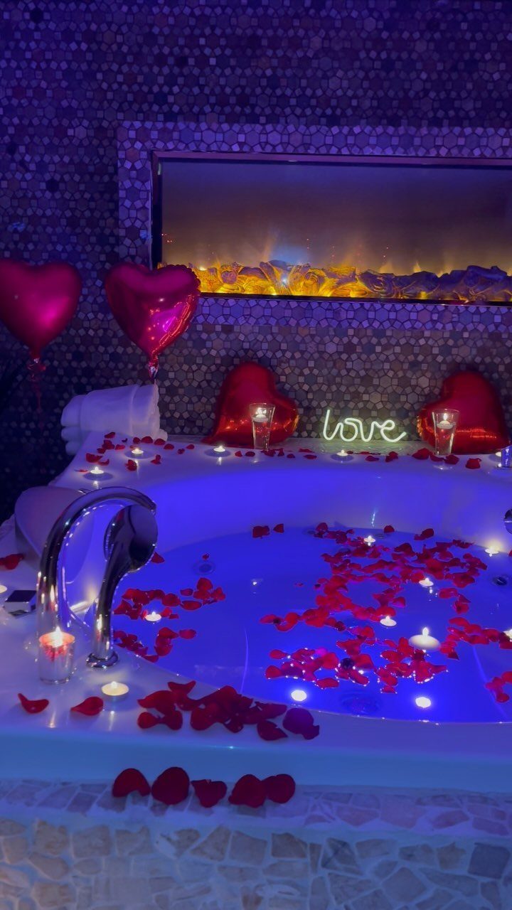 Romantic bathtub filled with rose petals, heart balloons, and candles under blue light.