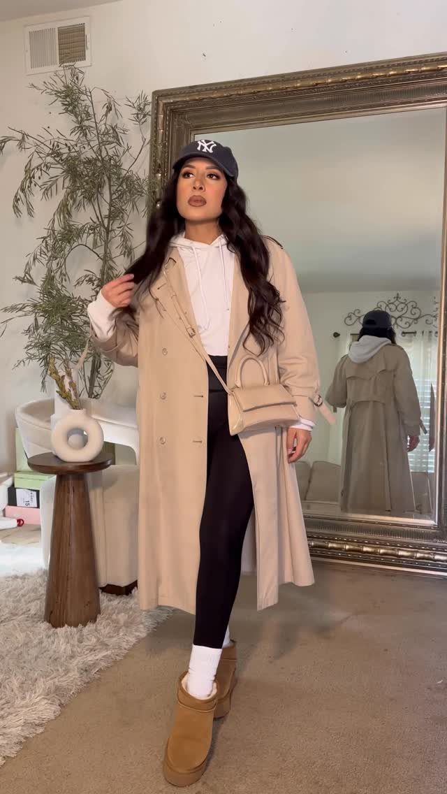 Woman in beige coat, white hoodie, and black leggings posing in a room.