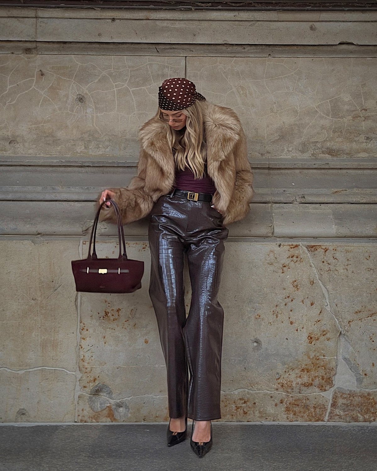 Woman in faux fur jacket and shiny brown trousers posing by stone wall